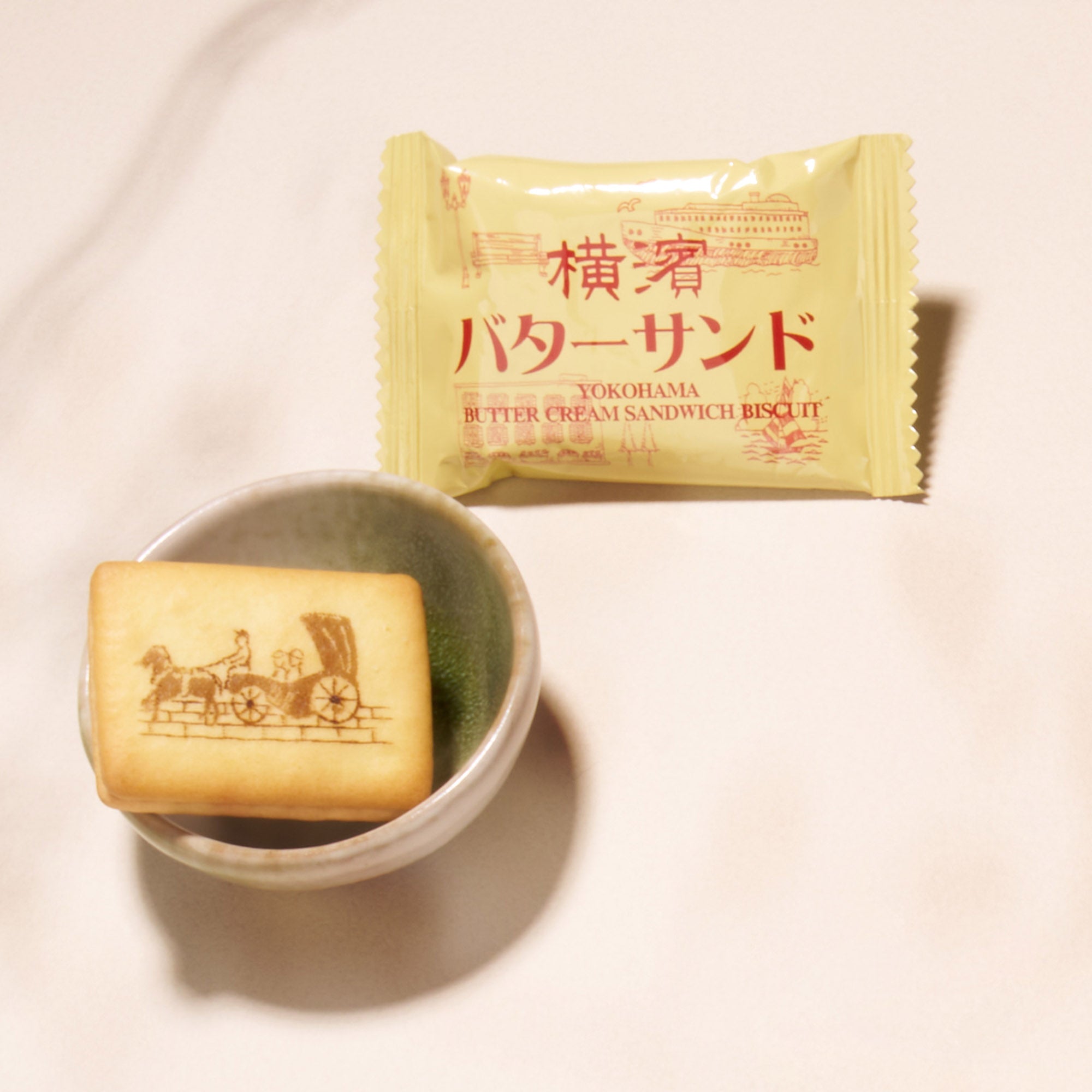 A butter cream sandwich biscuit with a carriage design sits in a bowl beside its yellow Japanese-English packaging—a delightful treat featured in the Bokksu Snack Box: Seasons of Japan.