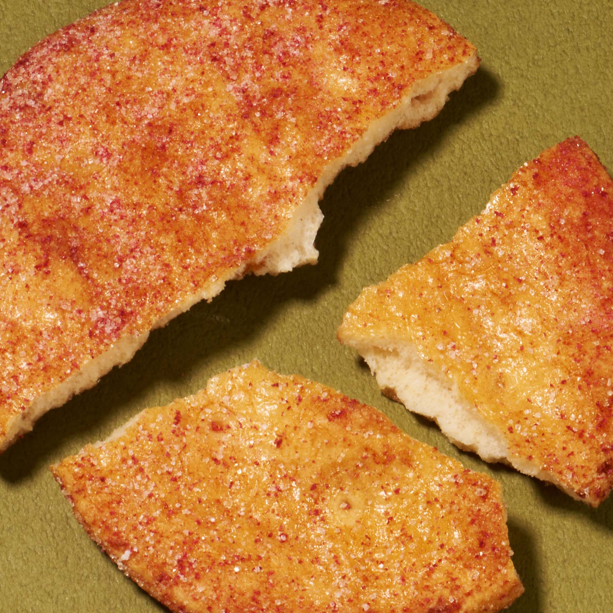 A round, golden-brown cookie topped with red sugar is broken into three pieces on a green surface, highlighting the delightful treats in the Bokksu Snack Box: Seasons of Japan.