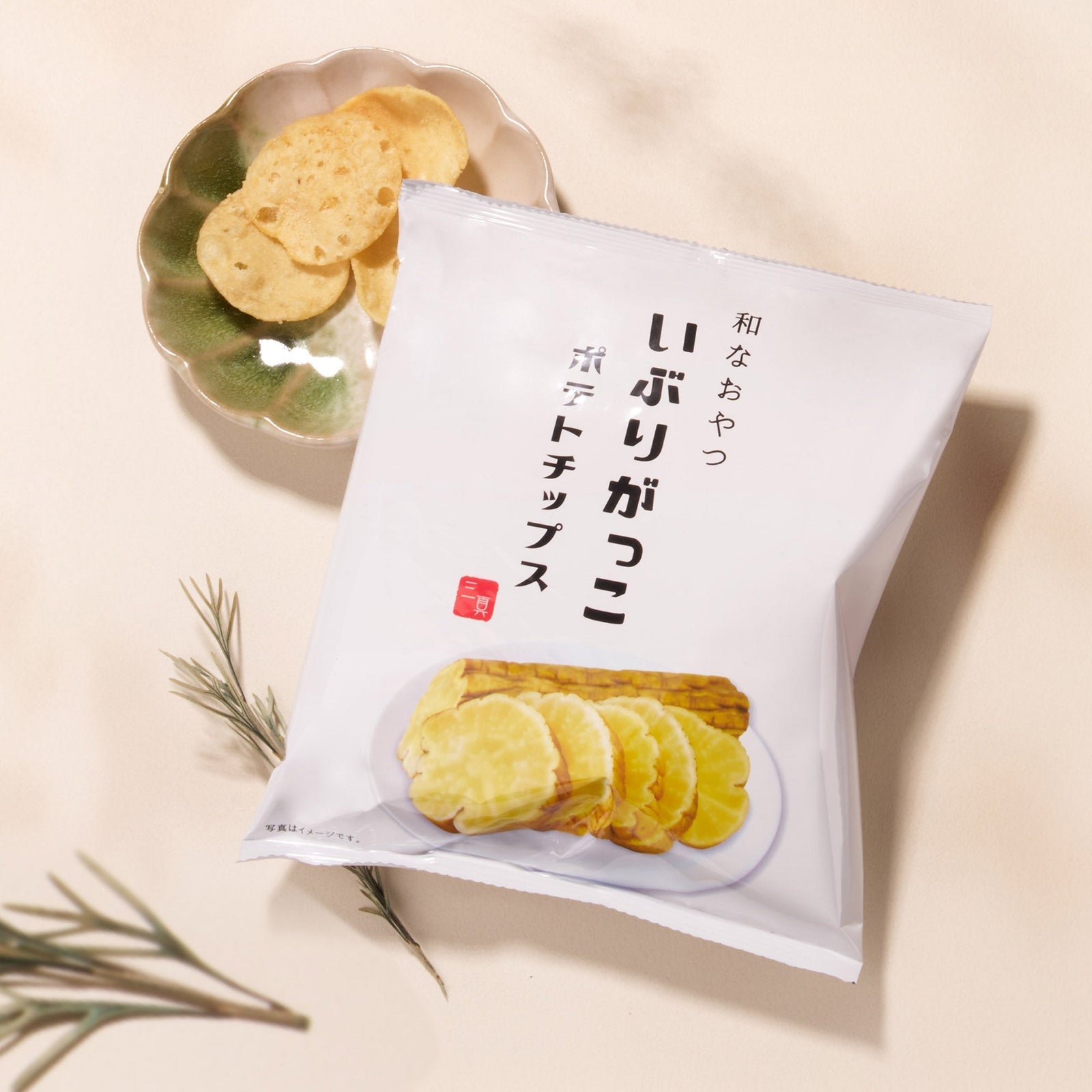 A Bokksu Snack Box: Seasons of Japan sits beside a small dish holding potato chips, with herb sprigs arranged nearby on a light background.